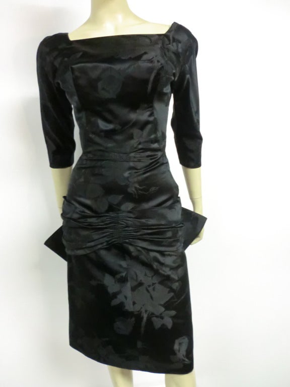 A fantastic 50s silk jacquard cocktail dress in black with a floral pattern:  zips up the back with a square neckline, 3/4 fitted sleeves, deep V back, a gathered swag rides low on the front hip and explodes in a swallowtail bow at back.  Custom