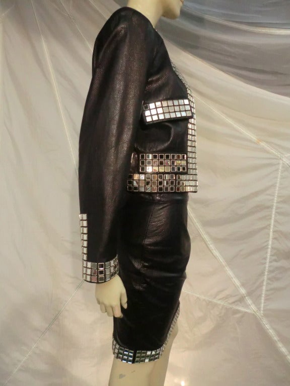 A Moschino black leather skirt suit that is a hybrid of Chanel style and biker attitude.  Trimmed with a wide band of mirror tiles at all edges, and pocket flaps, jacket is lined.  Jacket is a European size 42, the skirt is a 40.