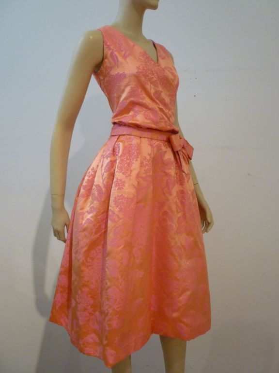 Un superbe original de Christian Dior datant de la fin des années 1950, en brocart floral corail et fuschia. Elle présente un corsage blouson de style enveloppant et une jupe ample froncée. La ceinture est façonnée et doublée de cuir de chevreau