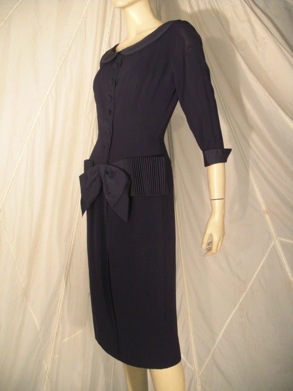 A beautifully tailored 1950s Bonwit Teller day dress:  Navy blue lightweight wool with self-covered buttons down front, a Peter Pan style collar and a wide cartridge pleated faille panel that ends in a bow at center front.  Turned up cuffs at the