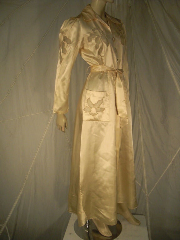 A darling 1940s pale peach satin lounging robe with sheer inset trimming on the bust, puff shoulders and pocket.  Slightly gathered waist and covered buttons. Matching belt included.