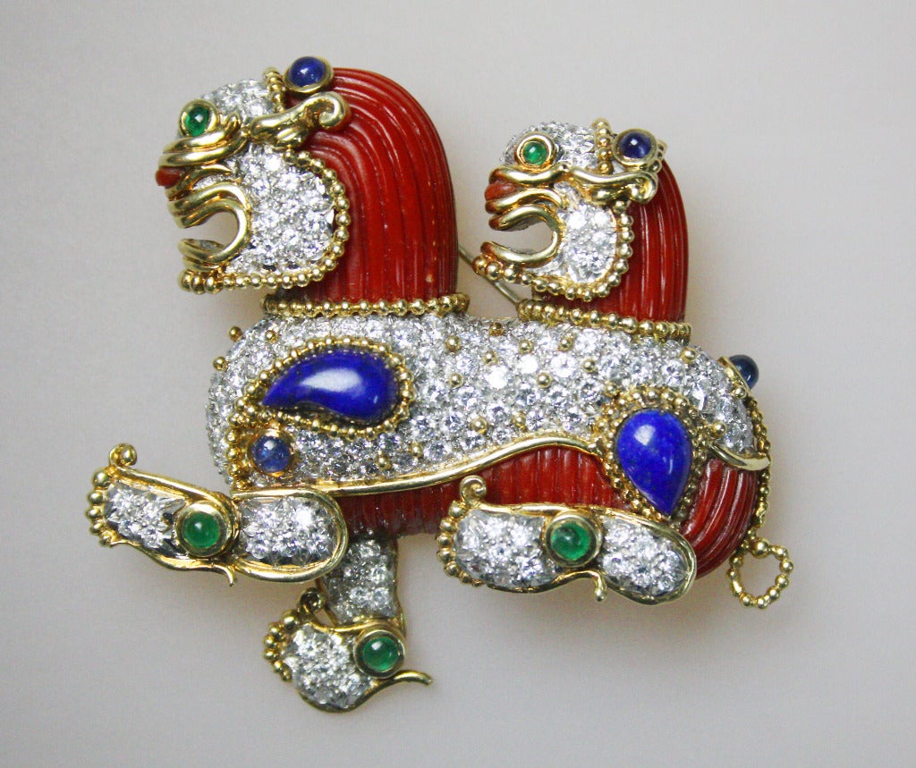 Modelled as a double headed lion dog know as Fo Dog , set with carved 'Ox Blood' coral, diamonds,  lapis lazuli,  emerald and sapphires cabochons. The brooch is 18K gold and is signed Cartier and is numbered.