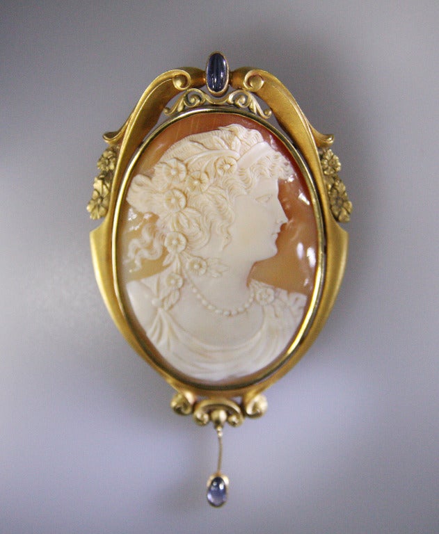 The cameo brooch was made in the early 1900s. It has a cabochon sapphire set in the top of the frame, and one hanging from the bottom of the frame. On the reverse side is a wire so that it can also be used as a pendant.