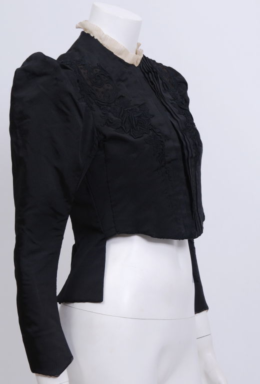 Form fitted black silk jacket with black silk applique design. Puffed shoulders, hook and eye closures, and cream colored  silk ruffled neckline.