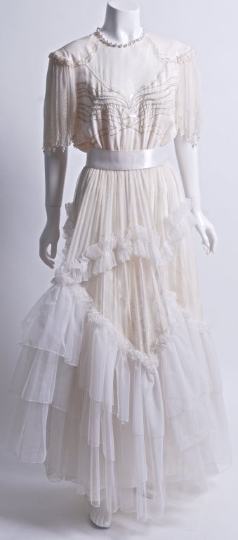 Rare, delicate silk chiffon, hand screened gown adorned with micro rhinestones, pearls and opalescent paillettes. Three tiers of silk net tulle, and scalloped ruffles. Very faint white on cream silk screened pattern throughout. Silk satin sash and