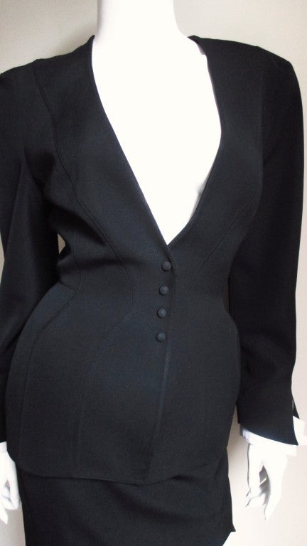 A fabulous black wool suiting skirt suit from Thierry Mugler. The collarless blazer has all of the piecing and curved seaming to create his signature hourglass shape. The cuffs and skirt hem have jagged hems with an additional layer shadowing the