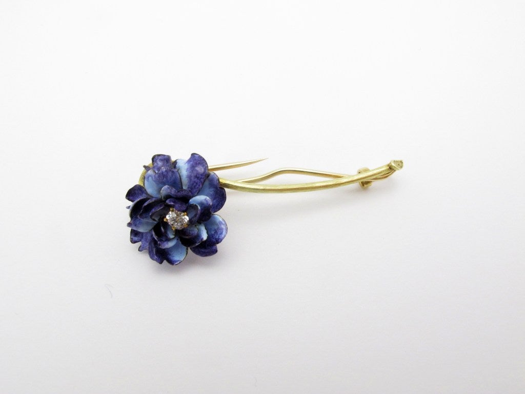 Signed Tiffany & Co. flower brooch. The petals are blue and lavender enamel set with a central old mine-cut diamond of approximate total weight 0.18ct, and the stem is lightly textured yellow gold.