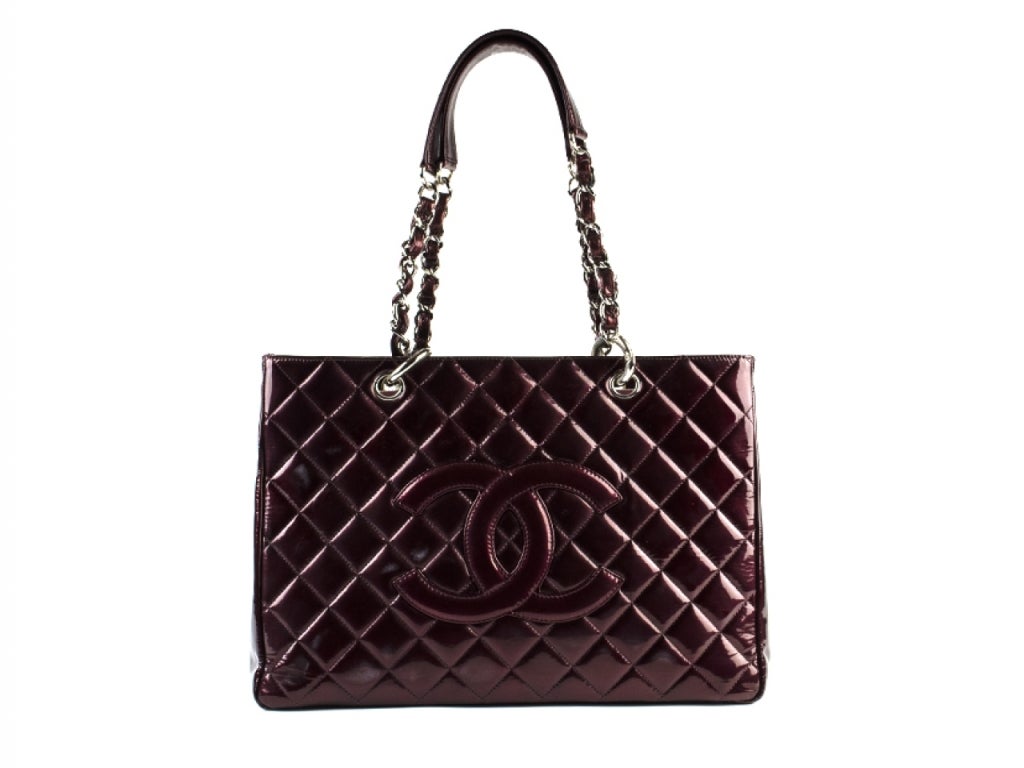 This stunning Chanel tote features shoulder straps with silver chain links threaded with patent leather shoulder pads as well as a prominent frontal patch Chanel CC logo and a rear pocket. The top zipper opens to a partitioned burgundy patent