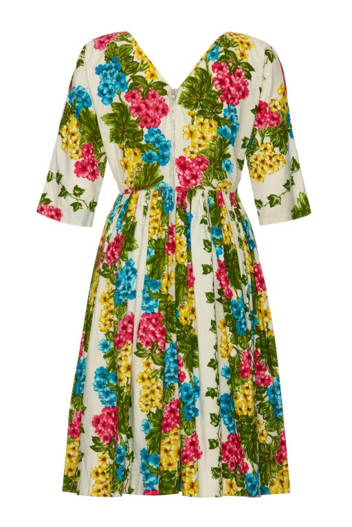 A great example of 1950s daywear, this fun cotton dress features a bold pink, green, blue and yellow floral print on a cream background. It has a full gathered skirt, ¾ sleeves and a deep V to the back and front neckline where there is also a pretty