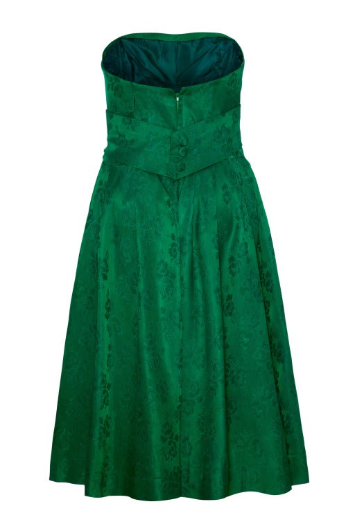 An adorable 50’s strapless cocktail dress in green silk jacquard with a woven floral pattern.
A pleated bust detail turns into a waistband that wraps around and fastens at the back with three large false covered buttons. 
There is boning around