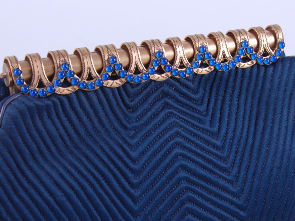 Navy blue satin evening bag from the art deco period with blue glass cabochon stones on the brass etched frame. The body of the bag is quilted in a zigzag pattern that cleverly joined on the base of the bag. The lining is dark blue satin.<br />
<br