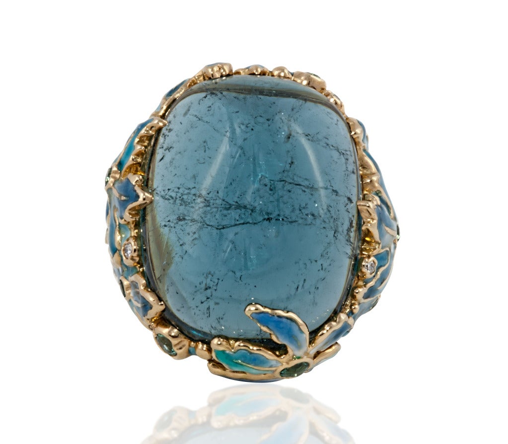 A piece unique, 18 karat yellow gold, hand-enameling, tourmaline and diamond ring.  The ring features a 61.11 carat, cabochon tourmaline set in 18 karat yellow gold and accented with hand-enameled flowers in blue hues.  The enameling is accented
