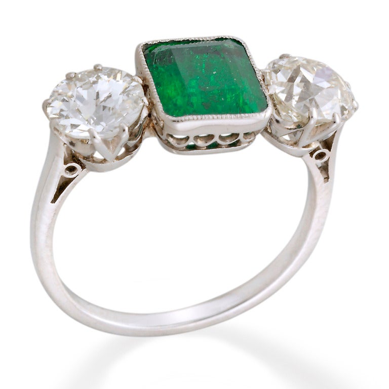 A mid 20th Century emerald and diamond three-stone ring, the central emerald-cut emerald estimated to weigh 1.5 carats, accompanied by GCS Report stating to be of Colombian origin, millegrain rubover-set to the centre to a palladium scalloped mount,