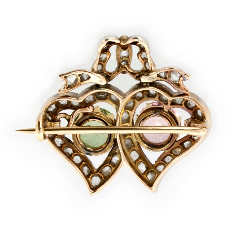 A late Victorian tourmaline and diamond twin heart brooch, the brooch set with a pink and green tourmaline, each estimated to weigh 1.25 carats, 8-claw set in yellow gold to the centre of a twin heart-shaped cluster surround with ribbon bow
