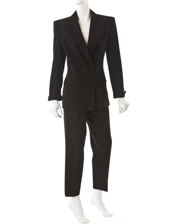 Black wool two-piece le Smoking pantsuit. Double-breasted blazer with a peak lapel, two front pockets and one breast pocket, hidden button closures, a double vent at back and folded cuffs. Pleated trousers have four pockets, a zip fly with hook