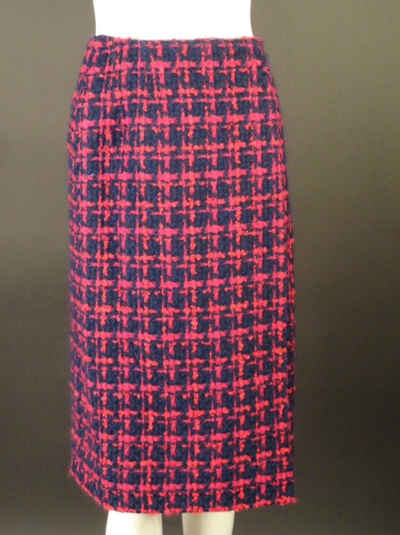 This timeless suit dates to the late 1980s. It is a fuchsia and navy wool tweed with a herringbone pattern.  The suit has a scoop neckline with a navy, loop ribbon fringe trim around that also runs down the center front. The same trim also edges the