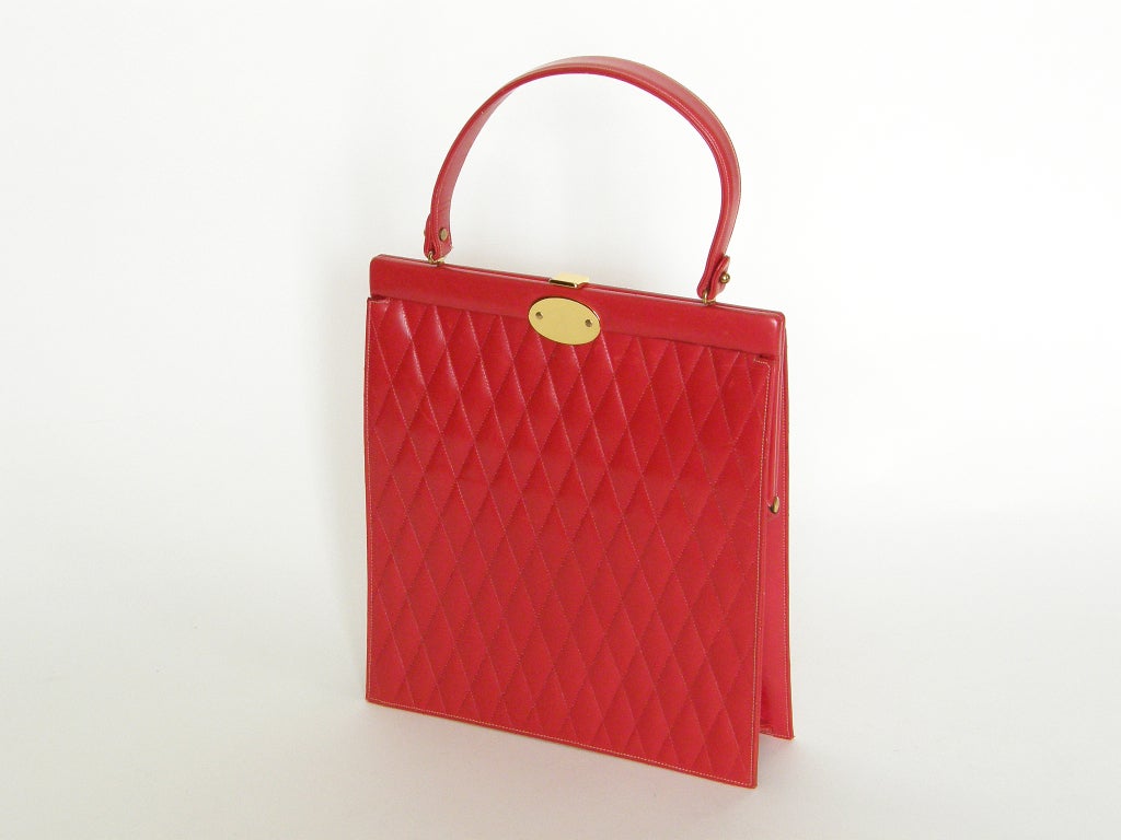 Red leather handbag by Prestige with a perfect balance of seduction and tasteful elegance. The bag has a slim and rigid form, and the body of the bag is quilted with a diamond shaped pattern. There is an oval plaque below the clasp for engraving.