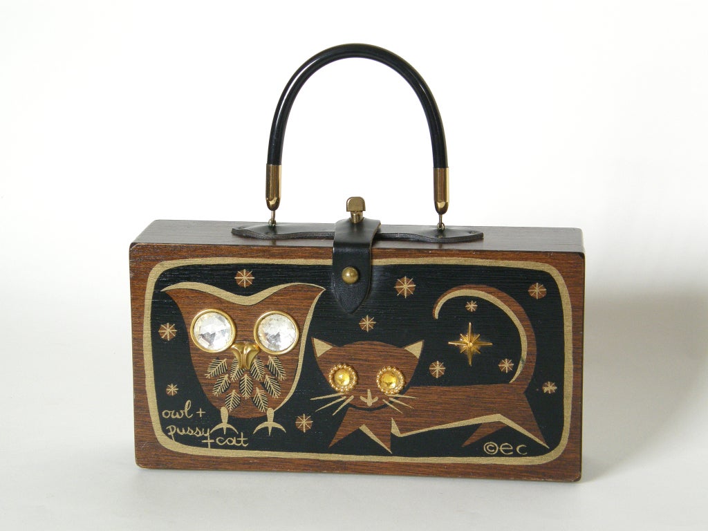 The owl and pussycat bag is one of Enid Collins classic designs. This wooden box bag has a rigid lucite handle and closes with a leather strap and brass toggle closure. The modernist, stylized owl and cat have large rhinestone eyes and gold metal