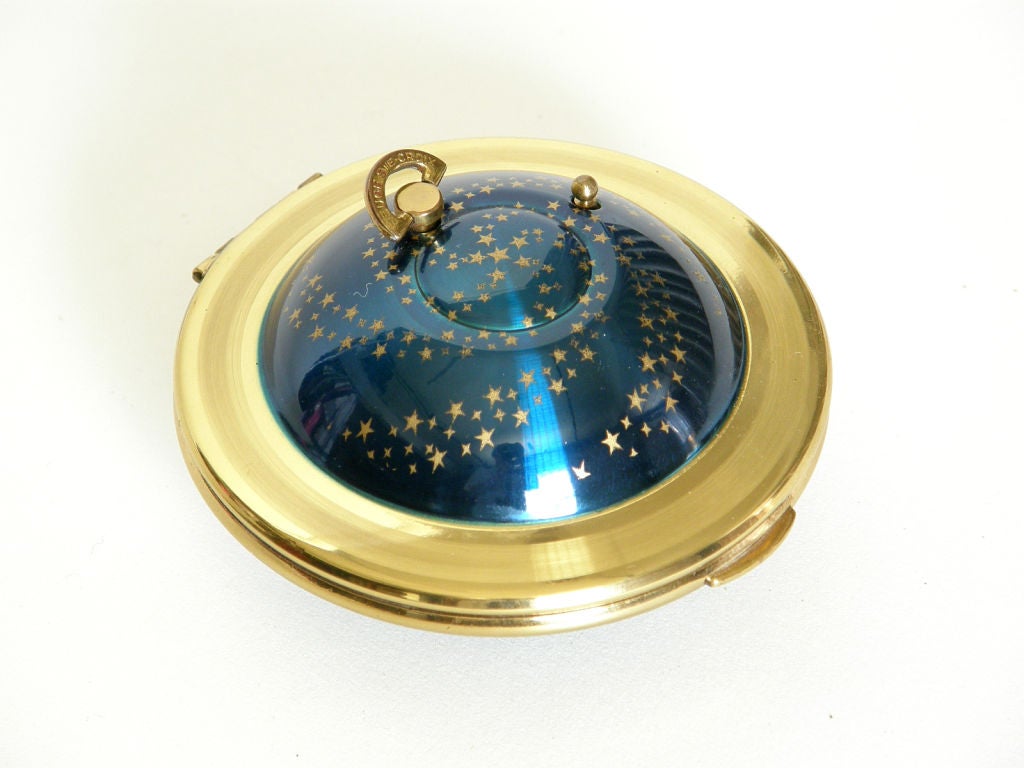 This fabulous flying saucer shaped compact has a built in music player. The attached key that winds it is on top, as is a small button that turn the musical function on and off. The domed parts are enameled with a swirling pattern of gold stars on a