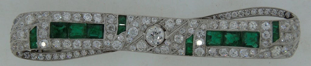 Classy and timeless Art Deco bow-tie pin created in Europe in the 1920's - 1930's. Features an Old European cut diamond in the center accented with additional diamonds and emeralds set in platinum. 
Old European cut diamond in the center weighs