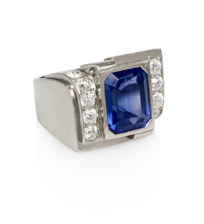An Art Deco sapphire and diamond ring of stylized geometric design with a central emerald-cut Ceylon sapphire, approximate total weight: 5.30 ct. (AGL certificate #CS 47140, no heat treatment), bordered on either side by a row of round brilliant