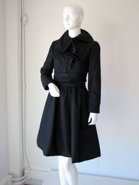 Fine vintage Geoffrey Beene cocktail dress. Black silk faille item features ruffle collar front (hidden snap closures), attached matching fabric wrap belt (hidden rear snap closures), full skirt & hidden side pockets.