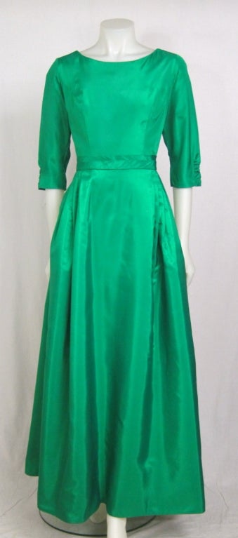 Featured is an emerald green taffeta gown from the early 1960s. Princess-seamed bodice with ruched, elbow length sleeves, and a double bias band at the waist, off-center pleats in the skirt. Low scooped back with flowing Watteau pleated train. Lined