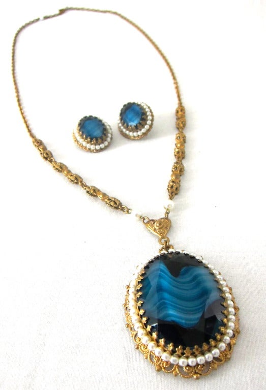 Featured is a lovely matched set of  clip on earrings and a necklace from West Germany. The Victorian style can be seen through the use of intricate brass filigree links, faux pearls, and decorative prongs. The blue glass is faceted and has white