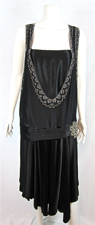Featured is a stunning vintage 1920s-era evening dress in fluid black satin and sparkling beadwork and rhinestones. The front and back neckline and armholes are richly embellished and the pleated hip detail also boasts a lovely rhinestone applique