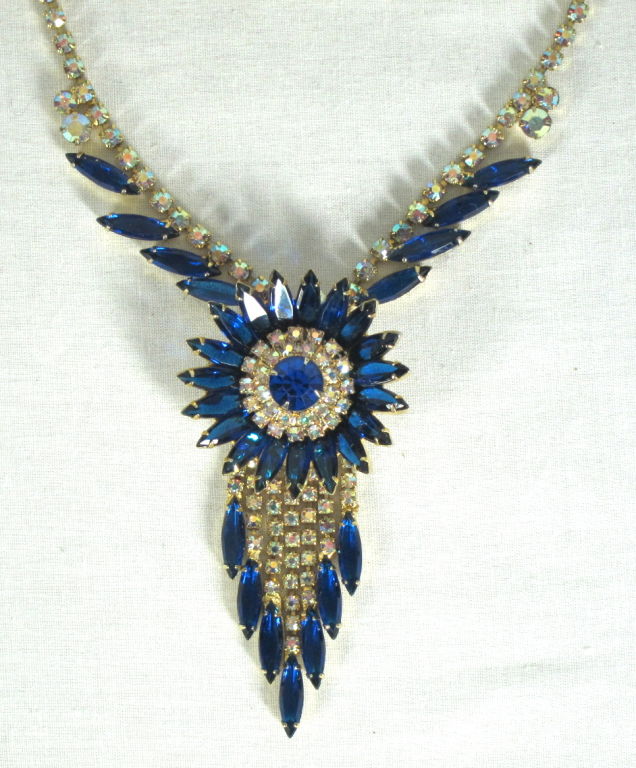 Featured is a stunning rhinestone necklace. A graduated fringe of 7 strands dangles underneath, each end.ing with a sapphire marquis stone. All stones are prong set. The base is a bright gold tone and the motif has a filigree backing. Closes with a