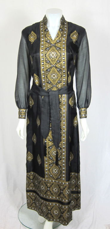Featured is a gorgeous hand printed Alfred Shaheen gown. It has a notched Mandarin collar, sheer sleeves, and allover gold and white paisley print. It is fully lined and fastens with a nylon back zipper. Includes the matching tasseled belt. <br