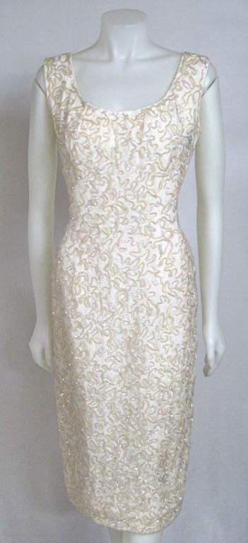 Featured is a sexy wiggle cocktail dress from the early 1960s. The fabric is ivory lace covered in meandering iridescent sequins, you are sure to shine in this dress! It is fully lined and fastens with a metal back zipper. From 