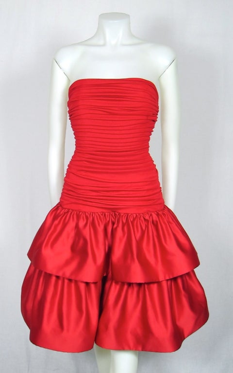 Featured is a flirty red party dress by Morton Myles. The fitted bodice is entirely ruched in fine jersey fabric and molds into a dropped waist. Twin tiers of matte satin and a back bow make up the bouffant skirt. Fully lined and fastens with a