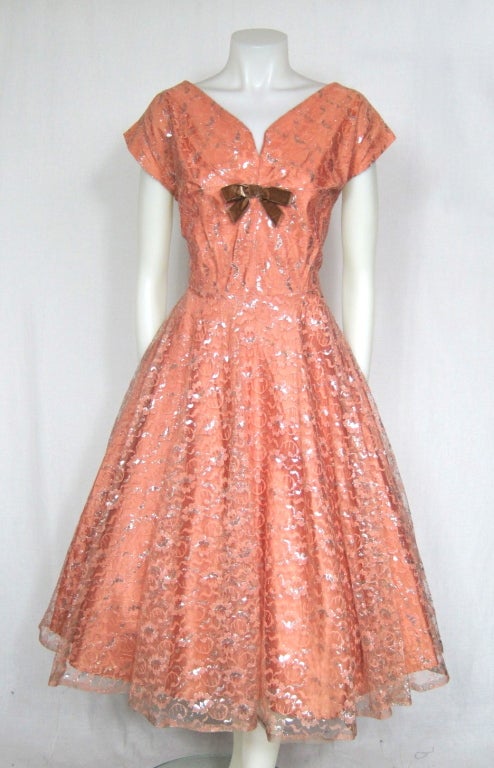 Featured is a lovely vintage party dress from the 1950s. It is made of a muted dusty pink floral lace with silver accents throughout. The fitted bodice has short, kimono-style sleeves, notched neckline and a velvet bow. The skirt is very full and is