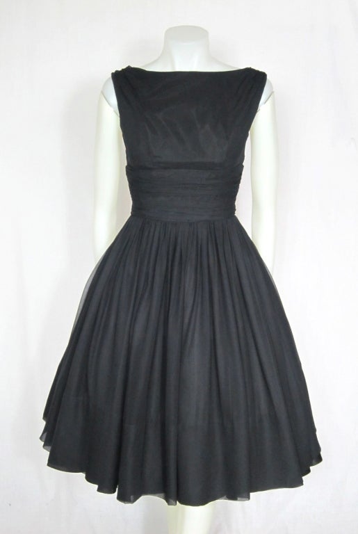 Featured is a dramatic black vintage cocktail party dress by Jr. Theme, from the 1950s. It has a bateau neckline, ruched midriff, and extremely full skirt. The back dips down and floating sashes extend from the shoulders, which can be arranged a