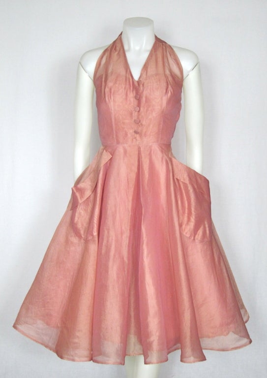 Featured is a gorgeous vintage party dress from the 1950s. Made of rose pink silk organza, it has a halter bodice with decorative buttons. The circle skirt has an inverted pleat in front and back and large front pockets. Nylon horsehair shapes the