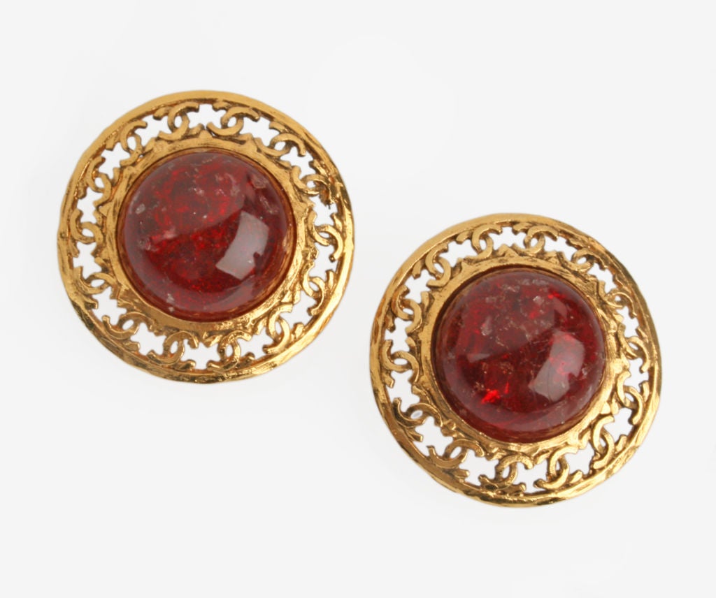 These are handsome CHANEL earrings with large poured red glass cabochons surrounded by a border of cutout CHANEL logos.  The glass measures .77