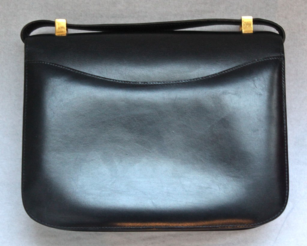 Navy blue box leather Constance bag with gold hardware from Hermes. Bag measures 9” wide, 6.5” tall, and expandable up to 2” deep. It has two inner compartments, with a zippered pocket. Exterior pocket at back. The strap can be worn long off your