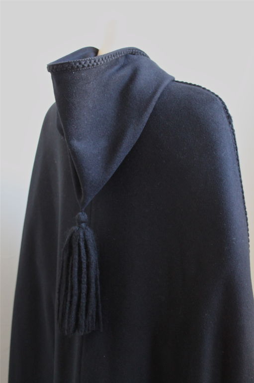 Dramatic floor length cape from Dior. This piece dates to the 1970's. It is jet black in color and trimmed with braiding,  which wraps around the center front and edges of the hood. The hood is also adorned by a long thick tassel. The side seams are