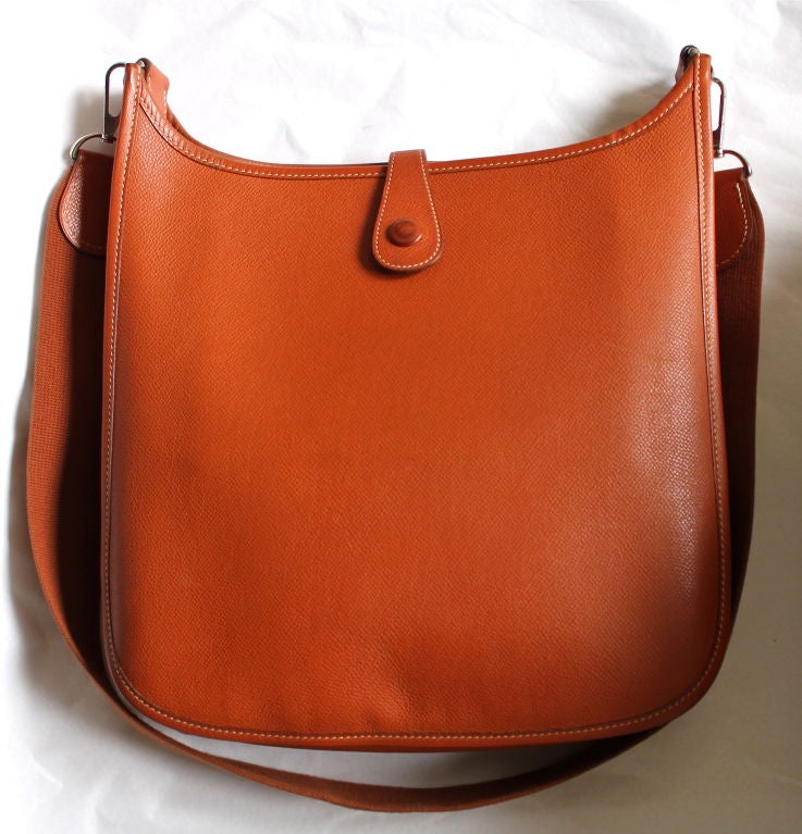 Classic 'Eveylyn' messenger style bag from Hermes. Sienna orange leather with palladium plated hardware. Bag measures approximately 12.5” x 11.5” x 3”. Canvas strap measures approximately 18” long. Unlined with one interior slot pocket. Made in