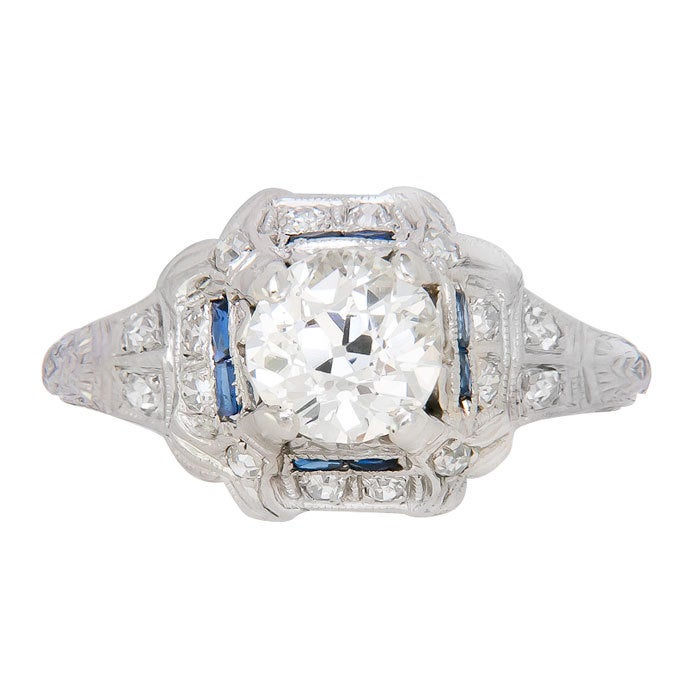 Circa: 1920s Platinum Engagement Ring, centrally set with a 1.30 Carat European cut Diamond that is L in Color and SI2 in Clarity, further surrounded by numerous smaller Diamonds and Sapphires, finished with Hand Engraved design work. Finger Size = 7