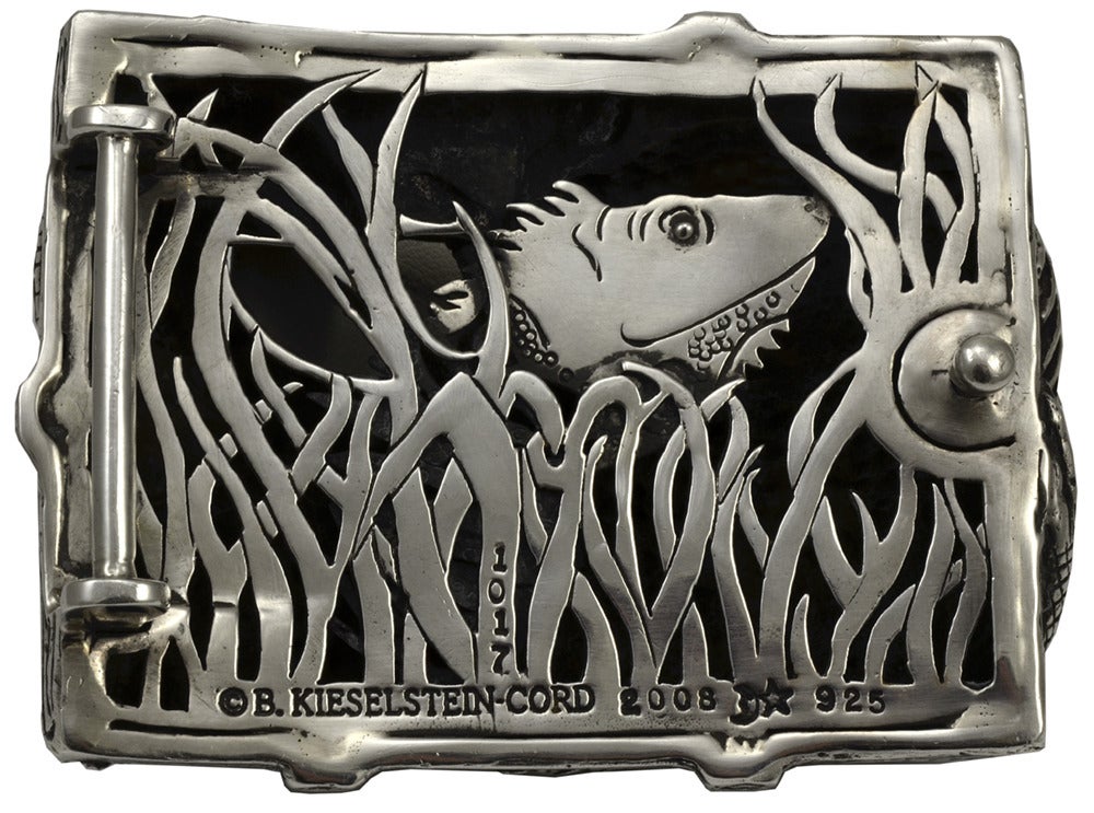 Very dramatic Sterling Silver belt buckle in the shape of an iguana perched on a log. Made, signed and numbered by B. Kieselstein- Cord. Very realistic life-like detailing; high relief gives a three-dimensional effect.  Glass eye.  Reverse side