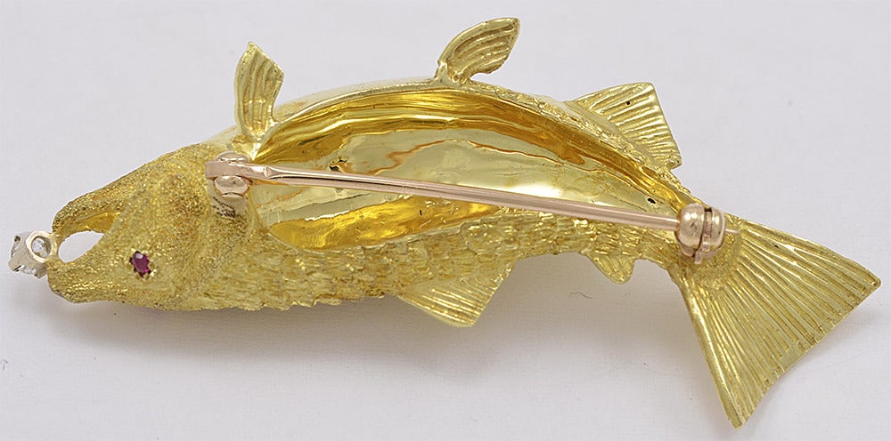 Beautifully detailed 18kt gold figural fish brooch. Faceted ruby eye.

Outstanding gold work which creates an all over shimmering texture. This fish is so lifelike you expect him to swim away with the diamond in his mouth.