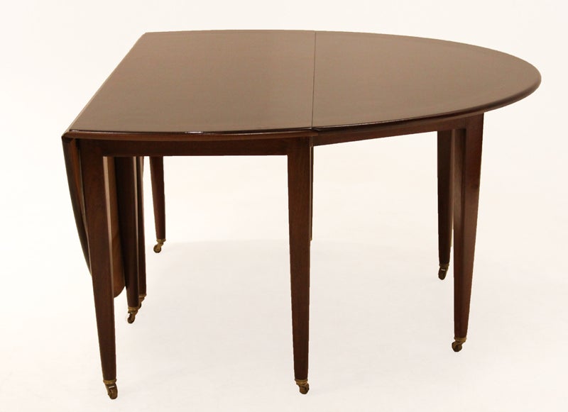 Oval drop-leaf Mahogany table by Dunbar. The table opens to seat eight and stimulates talk and simplifies informal dining, folding narrowly against a wall. The table has eight tapered legs with wheels, four or which are adjustable and fold in when