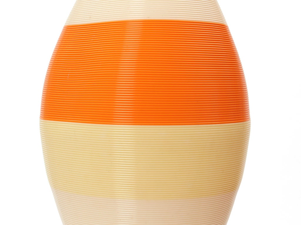 Una lampada a sospensione realizzata con corde di plastica continue che si intrecciano in un apparecchio da soffitto a forma di cono. I cavi bianco sporco, giallo e arancione producono una calda luce quando vengono accesi.