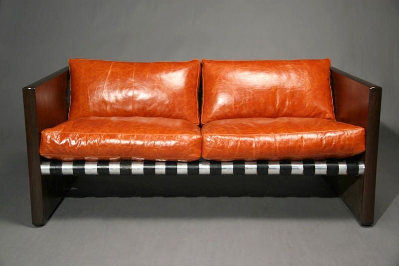 A sling sofa with black leather supports upholstered in orange leather with rosewood sides and chrome support poles.
Seat depth measures 19

Many pieces are stored in our warehouse, so please click on CONTACT DEALER under our logo below to find out