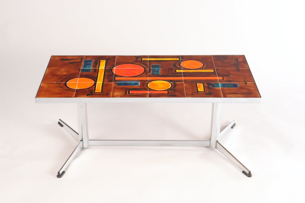 A petite, low-profile cocktail table with a polished chrome frame and a ceramic tile top featuring a geometric design in ultra-saturated shades of orange, yellow and blue with black outline on a mahogany ground. This is one of the nicest ceramic-top
