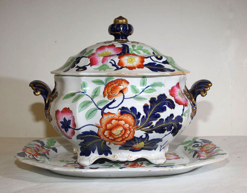 OF SHAPED OVAL FORM, WITH VOLUTE HANDLES, DOMED COVER WITH KNOB FINIAL AND ASSOCIATED OCTOGONAL UNDERPLATE, PAINTED WITH FLOWERS AND LEAVES IN COLOR ENAMELS, THE COBALT BLUE FINIAL AND HANDLES HIGHLIGHTED WITH GILT, WITH IMPRESSED MARK 