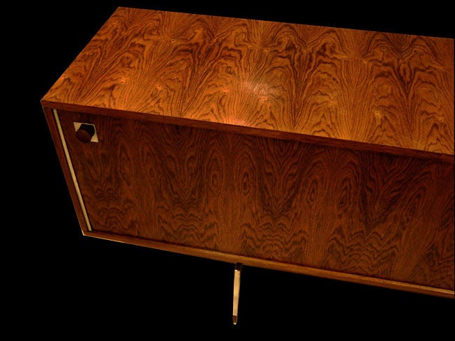 A long sideboard featuring a body in stained oak with two sliding doors, one in wood and one with an abstract modernist painting on tile of dancing figures. The sideboard sits on a very simple black steel frame with four chromed steel legs. By