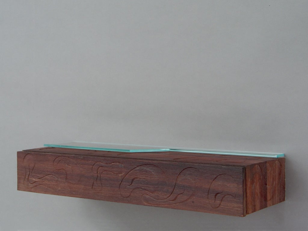 A rare and important wall mounted shelf build from solid mahogany. The rectangular shaped case holding one drawer and carrying an asymmetrical shaped glass shelf. The brushed surface showing carved interlocking abstracts elements. <br />
<br